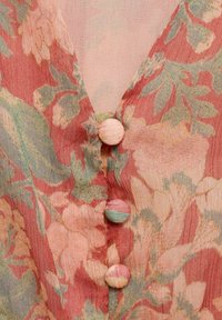 Primo piano di un tessuto floreale con tonalità rosa, verde e beige, caratterizzato da tre bottoni rivestiti in tessuto lungo un scollo a V.