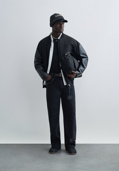 Veste bomber noire avec manches en cuir portée sur un t-shirt noir, pantalon en denim déchiré avec des embellissements argentés, et une casquette à motif.