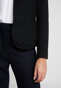 Černý blazer s texturovaným povrchem, zdobený malými bílými tečkami, s jedním knoflíkem a kapsou. Nosený přes bílé tričko a tmavé kalhoty.