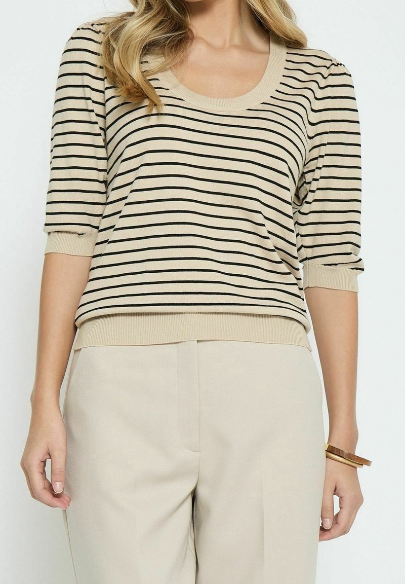 Femme portant un pull beige à manches courtes avec des rayures horizontales noires et un pantalon beige, accessoirisée avec des bracelets en bois au poignet droit.