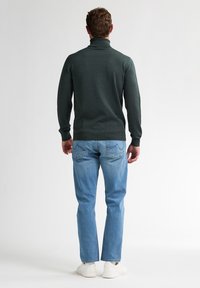 Pulover verde închis cu guler înalt, realizat dintr-un material moale, purtat cu blugi albaștri deschiși și adidași albi, văzut din spate.