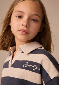 Gestreepte polo in beige en marineblauw, met een platte kraag en geborduurd "Sports Club" logo op de voorkant. Getextureerd brei-materiaal.