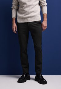 Ljusgrå stickad tröja med ribbad fåll, matchad med svarta slim-fit byxor och kraftiga svarta snörskor. En enfärgad blå bakgrund.