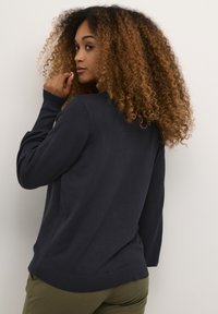 Tumši jūras zila džemperis ar nedaudz brīvu piegriezumu, ribotām piedurknēm un noapaļotu apakšmalu. Mīksta faktūra ar minimāla apdare aizmugurē.