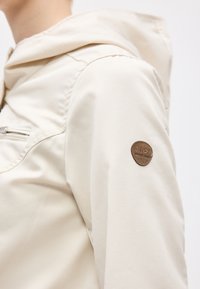 Μανίκι μπεζ σακακιού με ένα μικρό στρογγυλό καφέ έμπλαστρο στο πάνω μέρος του βραχίονα και μια θήκη στο στήθος με φερμουάρ στο σώμα.