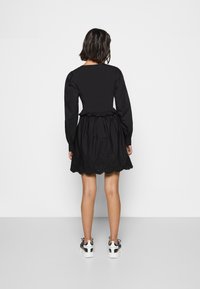 Vestido preto com mangas bufantes longas, corpete ajustado e saia evasé com bainha de ilhós em forma de onda. Usado com ténis brancos e pretos.