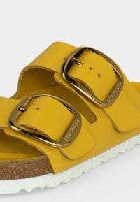 Κίτρινο σανδάλι Birkenstock με δύο μεγάλες χρυσές αγκράφες και φελλό πάτο με λευκή σόλα, εμφανίζεται σε κοντινή γωνία.