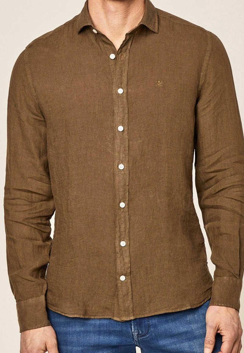 Homme portant une chemise marron à manches longues avec des boutons blancs et un jean bleu, visible du cou aux hanches.