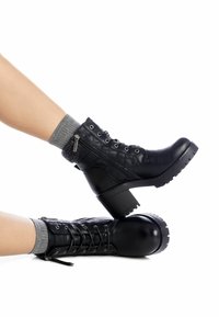 Botas negras hasta el tobillo con un patrón acolchado, diseño de cordones, cremallera lateral y tacón grueso. Calcetines grises visibles por encima del collar de la bota.