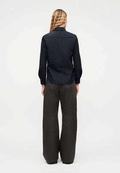 Femme aux longs cheveux blonds portant une chemise noire boutonnée et un pantalon large gris foncé, debout de dos devant un fond uni.