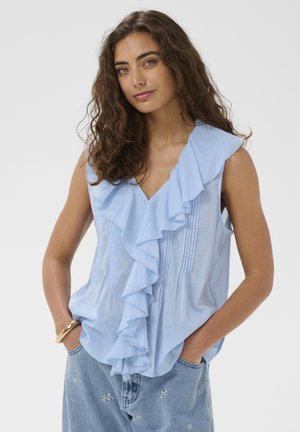 Vrouw met krullend bruin haar, draagt een mouwloze lichtblauwe blouse met ruches en spijkerbroek, handen in de zakken, neutrale achtergrond.