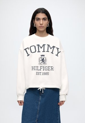 Jeune femme aux longs cheveux foncés porte un sweat-shirt blanc Tommy Hilfiger avec logo et une jupe en jean bleue, debout devant un fond uni.