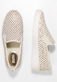 Chaussures beige à enfiler avec une tige en toile, présentant des découpes en forme d'étoiles dorées, une semelle blanche épaisse et une semelle intérieure métallique dorée avec marquage.