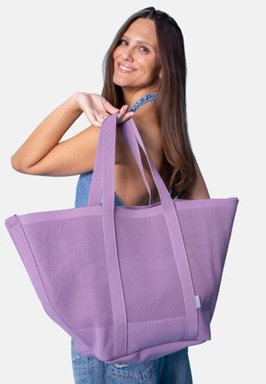 Smiling woman in denim overalls holding a large lavender mesh tote bag over her shoulder against a plain background.