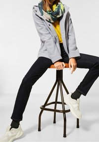 Grå fleecejacka, gul topp, mörka jeans och vita sneakers. Halsduk med grön och beige mönster. Sitter på en träpall.