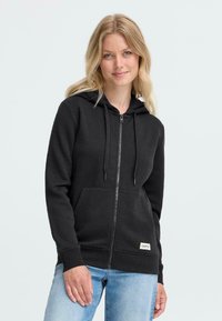 Svart zip-up hettegenser med snor i hetta, frontlommer, ribbestrikkede mansjetter, og et lite hvitt logopatch nederst. Mykt, teksturert stoff.