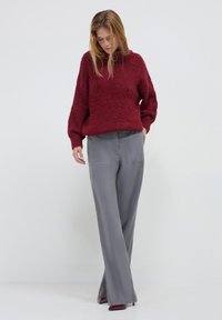 Donna che indossa un maglione borgogna strutturato e pantaloni grigi a gamba larga, in piedi con una mano in tasca e lo sguardo rivolto verso il basso.