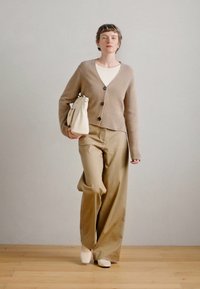 Cardigã de malha bege com botões escuros, blusa branca, calças largas bege e calçado branco. Agarra uma mala de cor creme. Linhas simples e limpas.