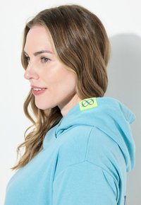 Felpa azzurra chiara con una vestibilità rilassata, caratterizzata da una tasca a marsupio frontale e un vivace accento del logo verde sulla spalla.