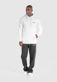 Sweat-shirt à capuche blanche avec poche avant et logo "SMILODON", assorti à un pantalon de survêtement gris foncé portant le même logo, porté avec des baskets blanches.