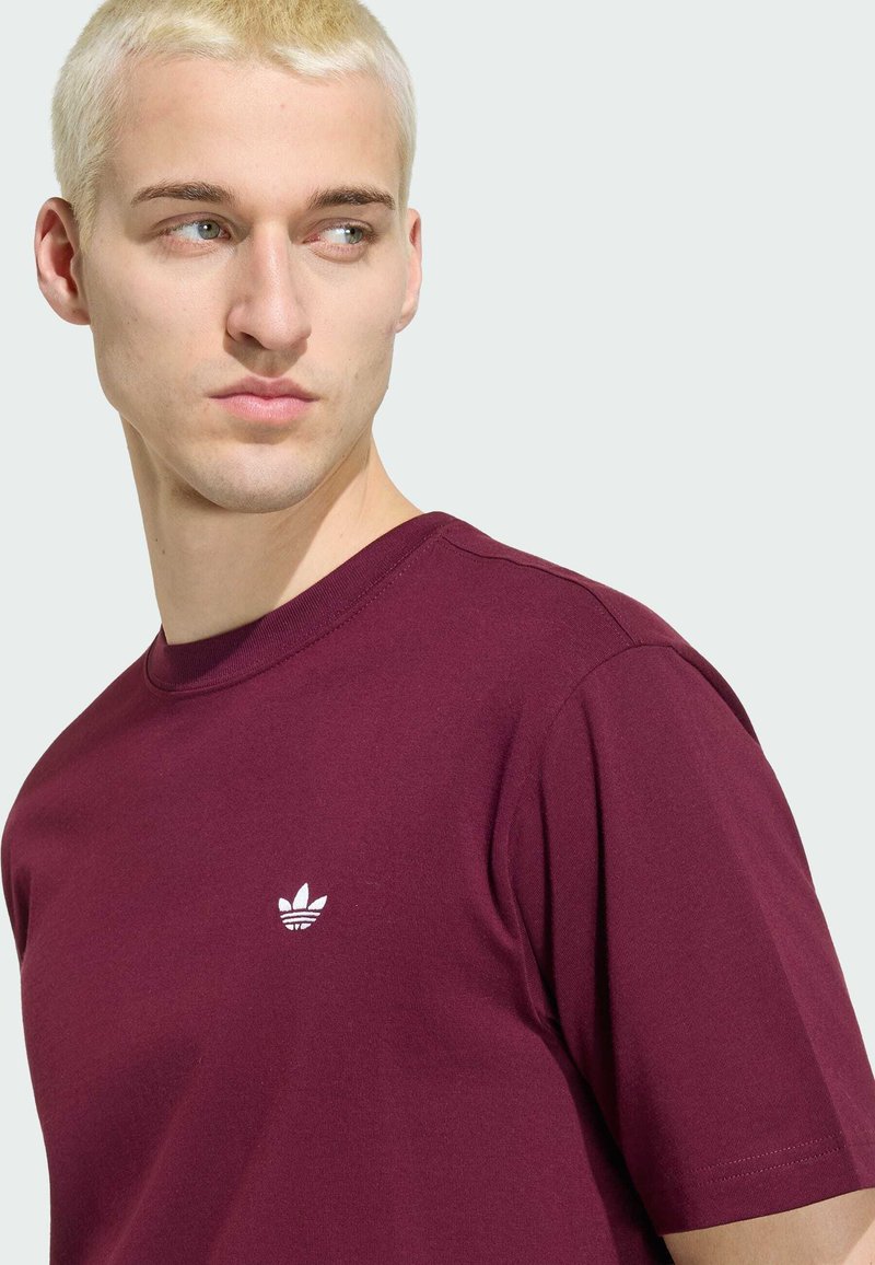 Lyshåret mand, der kigger mod venstre, iført en ensfarvet mørkerød Adidas T-shirt med et lille hvidt logo på brystet mod en lys baggrund.
