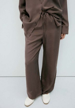 Personne portant un pantalon marron ample avec une chemise boutonnée assortie, les mains dans les poches, et des chaussures blanches pointues sur un sol clair.