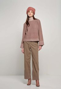 Overmaatse, geribbelde beige sweater gecombineerd met lichtbruine corduroy broek. Het model draagt een gemêleerde roze beanie en bruine haklaarzen.