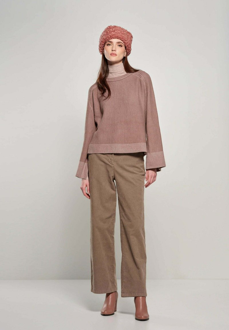 Overmaatse, geribbelde beige sweater gecombineerd met lichtbruine corduroy broek. Het model draagt een gemêleerde roze beanie en bruine haklaarzen.