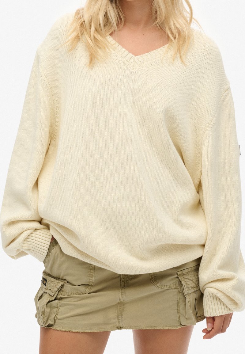 Personne portant un pull en maille crème oversize et une mini-jupe cargo vert olive avec des poches, debout devant un fond blanc.