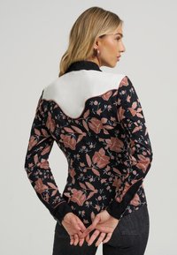 Blusa com padrão floral e base preta, apresentando pala branca estruturada e piping contrastante. Mangas compridas com punhos ajustados.