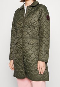 Olivgrön vadderad jacka med krage, dragkedjestängning och tryckknappsluckor. Har en cirkulär patch på vänster ärm och en slät yta.