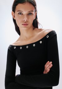 Fekete, vállakat szabadon hagyó felső hosszú ujjakkal, ezüstszínű fémkarikákkal díszítve a nyakkivágás mentén, sima, testhezálló anyagból készült.