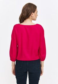 Fuksjowa bluzka z szerokim dekoltem i marszczonymi rękawami, wykonana z gładkiego materiału. Noszona z ciemnymi dżinsami, prezentującymi luźny krój.