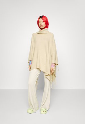 Poncho en tricot beige avec un col montant, des franges sur les côtés et une coupe ample ; associé à un pantalon crème à côtes et des sabots vert clair.
