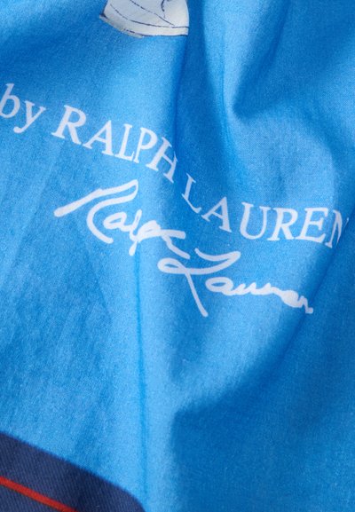Μπλε ύφασμα με λευκή επωνυμία «Ralph Lauren» και κείμενο υπογραφής, που δείχνει τις πτυχώσεις και την υφή του υλικού.