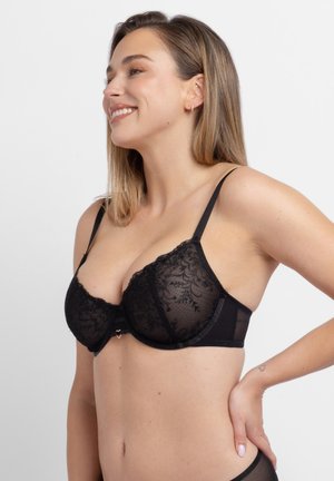 Uśmiechnięta kobieta z długimi włosami w czarnym koronkowym biustonoszu, pozująca z jedną ręką na biodrze na jednolitym tle.