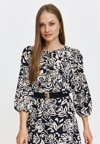 Luźna granatowa sukienka z beżowym kwiatowym wzorem, okrągły dekolt ze związanym wiązaniem, bufiaste rękawy, wypukła talia z czarnym paskiem i złotą klamrą.