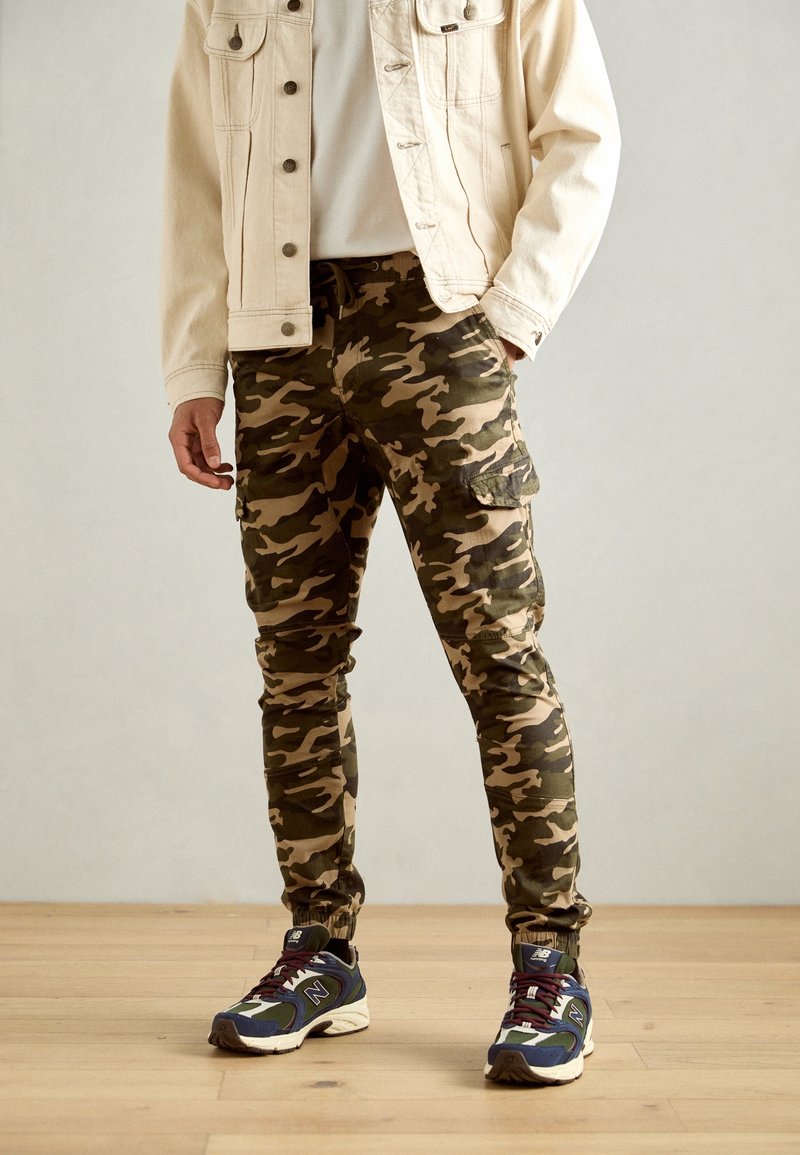 Personne portant un pantalon cargo camouflage, des baskets bleues et blanches, et une veste beige clair, debout sur un sol en bois.