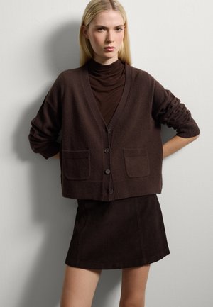 Cardigan marrone lavorato a maglia con scollatura a V, due tasche frontali e bottoni. Abbinato a una gonna a coste marrone coordinata. Texture liscia.