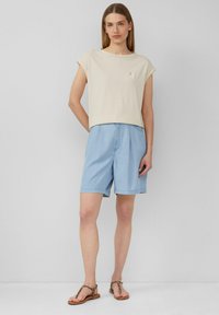 Beige katoenen t-shirt met opgerolde korte mouwen, gecombineerd met lichtblauwe denim korte broek. Kleurige sandalen met teenlus en textuuraccenten op de band.