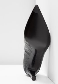 Chaussure à talon haut noire à bout pointu avec un motif texturé en peau de serpent, photographiée debout sur le talon contre un fond blanc en coin.
