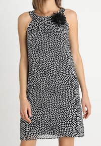 Mustavalkoinen pilkullinen mekko pyöreällä pääntiellä, kukkakoriste olkapäällä ja rento siluetti. Silkkinen, kevyt kangas.