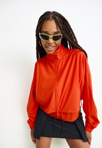 Lysende oransje, lett zip-up jakke med høy krage og elastiske mansjetter, kombinert med et svart miniskjørt. Solbriller med grønn kant.