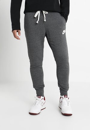 Personne portant un pantalon de survêtement Nike gris foncé avec des cordons blancs et des baskets Nike blanches avec des accents bordeaux, debout sur un sol blanc.