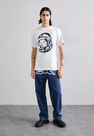 White cotton t-shirt featuring a navy astronaut graphic, paired with blue denim jeans and black slip-on shoes. Simple design, relaxed fit.