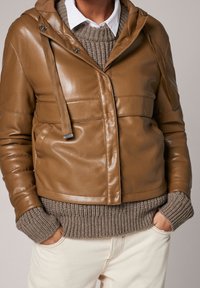 Veste en cuir brun avec une finition brillante, dotée d'une capuche, un pull en maille texturé en dessous et un pantalon couleur crème.