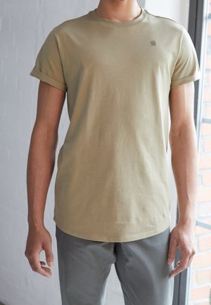 Man draagt een beige T-shirt met korte mouwen met opgerolde manchetten en lichtgrijze broek, staand binnen vlak bij een raam met baksteendetails.