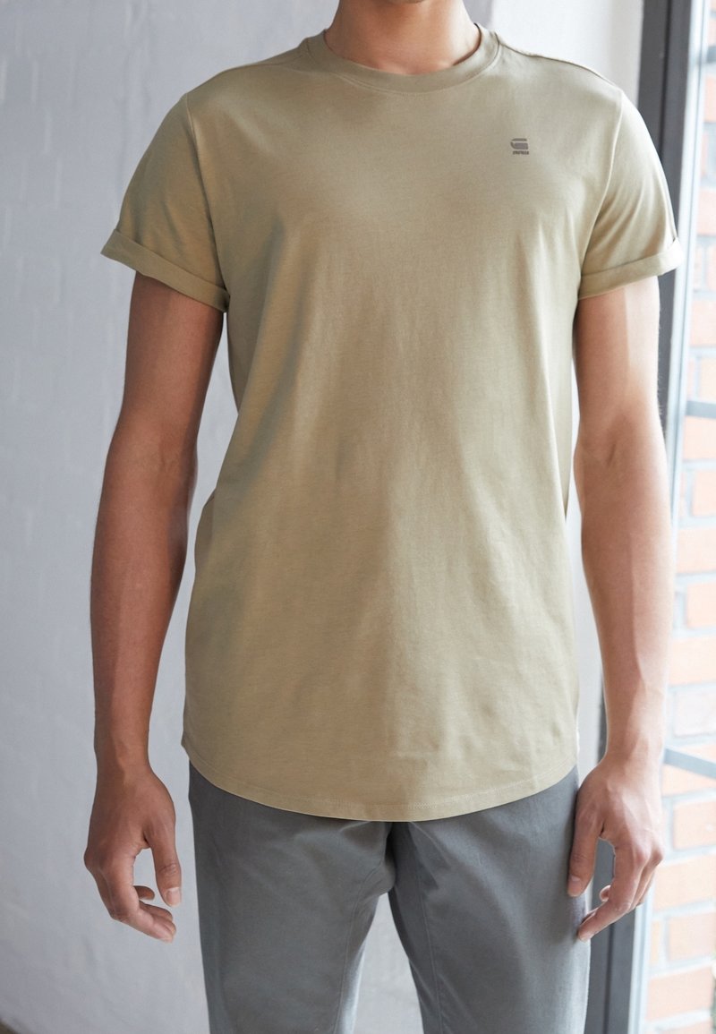 Homme portant un t-shirt beige à manches courtes avec les poignets retroussés et un pantalon gris clair, debout à l'intérieur près d'une fenêtre avec des détails en briques.