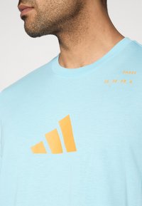 Ανοιχτό μπλε βαμβακερό μπλουζάκι με πορτοκαλί γραφικό τριών ρίγων στο μπροστινό μέρος και κείμενο που αναγράφει "PADEL 15_30_40_A." Στρογγυλή λαιμόκοψη.