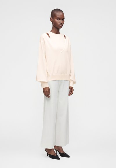 Femme debout portant un sweat-shirt crème avec des découpes aux épaules, un pantalon blanc à jambes larges et des chaussures à talons bas noires à bout pointu sur un fond uni.